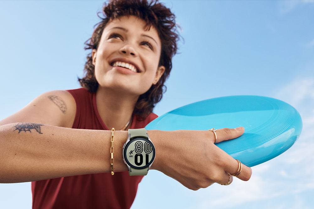 A woman is smiling and holding a frisbee while wearing a silver Galaxy Watch4 on her wrist.